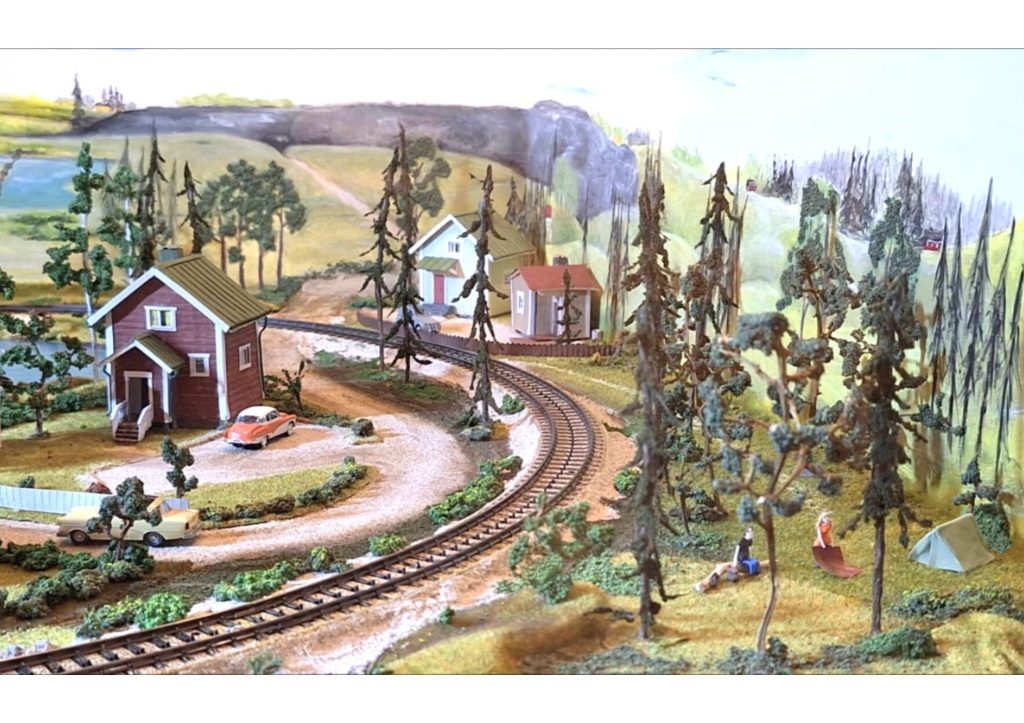 Diorama, jossa metsäisen junaradan varrella pieniä mökkejä, vanhoja autoja ja telttailijoita.