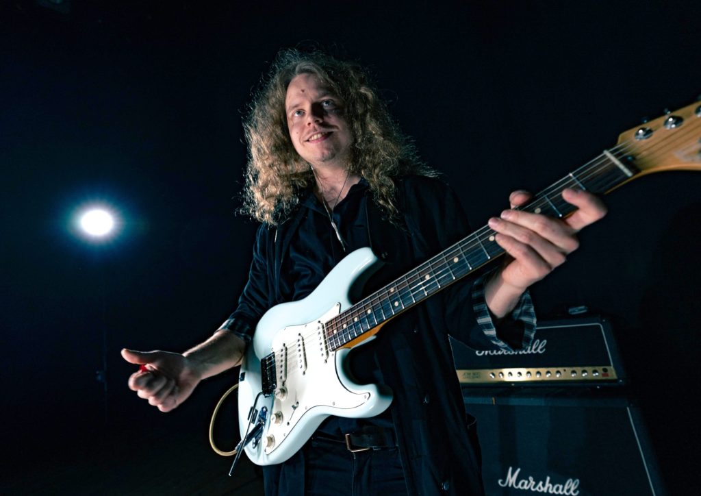 Tumma studiokuva Markus Venehsalosta valkoisen sähkökitaransa kanssa.