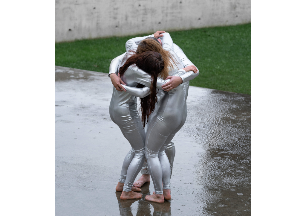 Kuva tanssiteatteri Minimin Alien-esityksestä: kolme hopeanvärisiin kokotrikoisiin pukeutunutta tanssijaa seisoo yhteen puristautuneina ulkona sateessa.